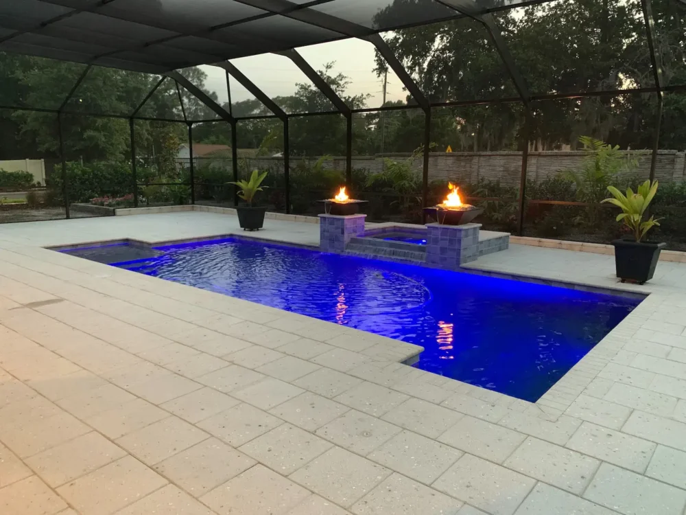 Dusk view of custom geometric pool with LED lighting, spillover spa, & fire bowls with water feature inside screened enclosure.