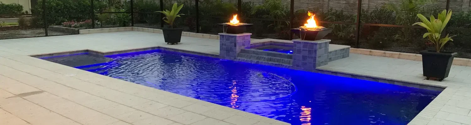 Dusk view of custom geometric pool with LED lighting, spillover spa, & fire bowls with water feature inside screened enclosure.