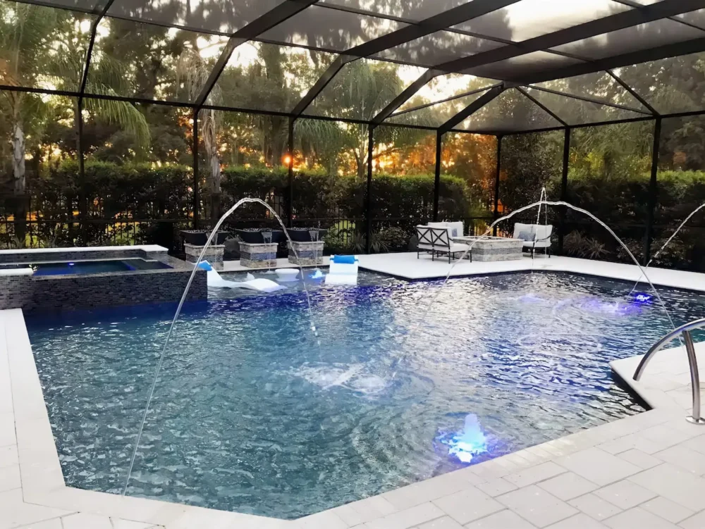 Geometric pool with spillover spa, multiple water features, fire bowls, sun shelf, LED lighting, & fire pit inside screen enclosure.