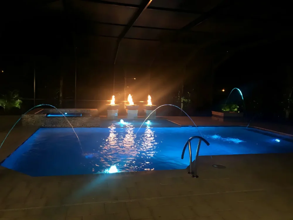 Night view of geometric pool with LED lighting, spillover spa, fire bowls, and multiple water features
